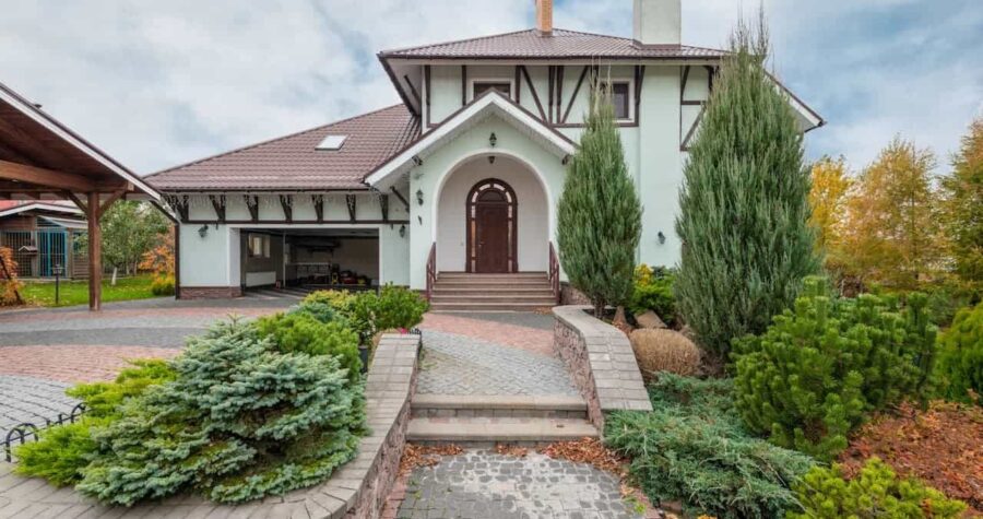 White Tudor-style house with arched entrance, landscaped pathway, evergreen shrubs, and cobblestone driveway under cloudy autumn sky