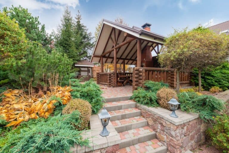 Rustic wooden gazebo with log railings accessed by brick steps, surrounded by evergreen shrubs and autumn foliage