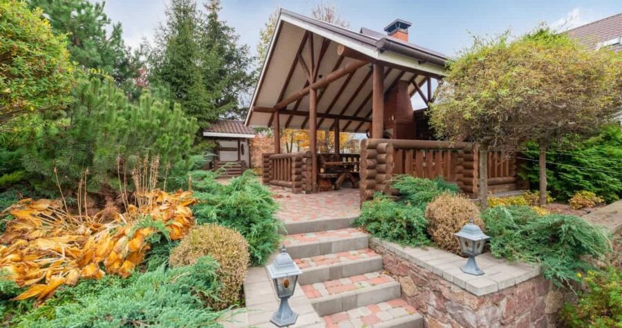 Rustic wooden gazebo with log railings accessed by brick steps, surrounded by evergreen shrubs and autumn foliage