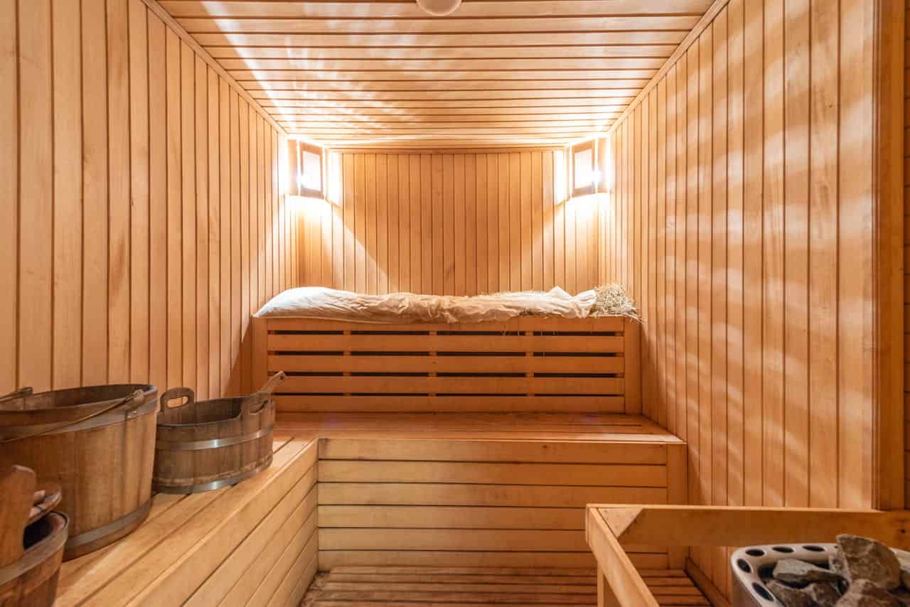 Wooden sauna room with light wood paneling, a wooden bench, small wooden buckets for sauna use, a pile of hay on the upper shelf, soft lighting, and a relaxing atmosphere, designed for relaxation and wellness