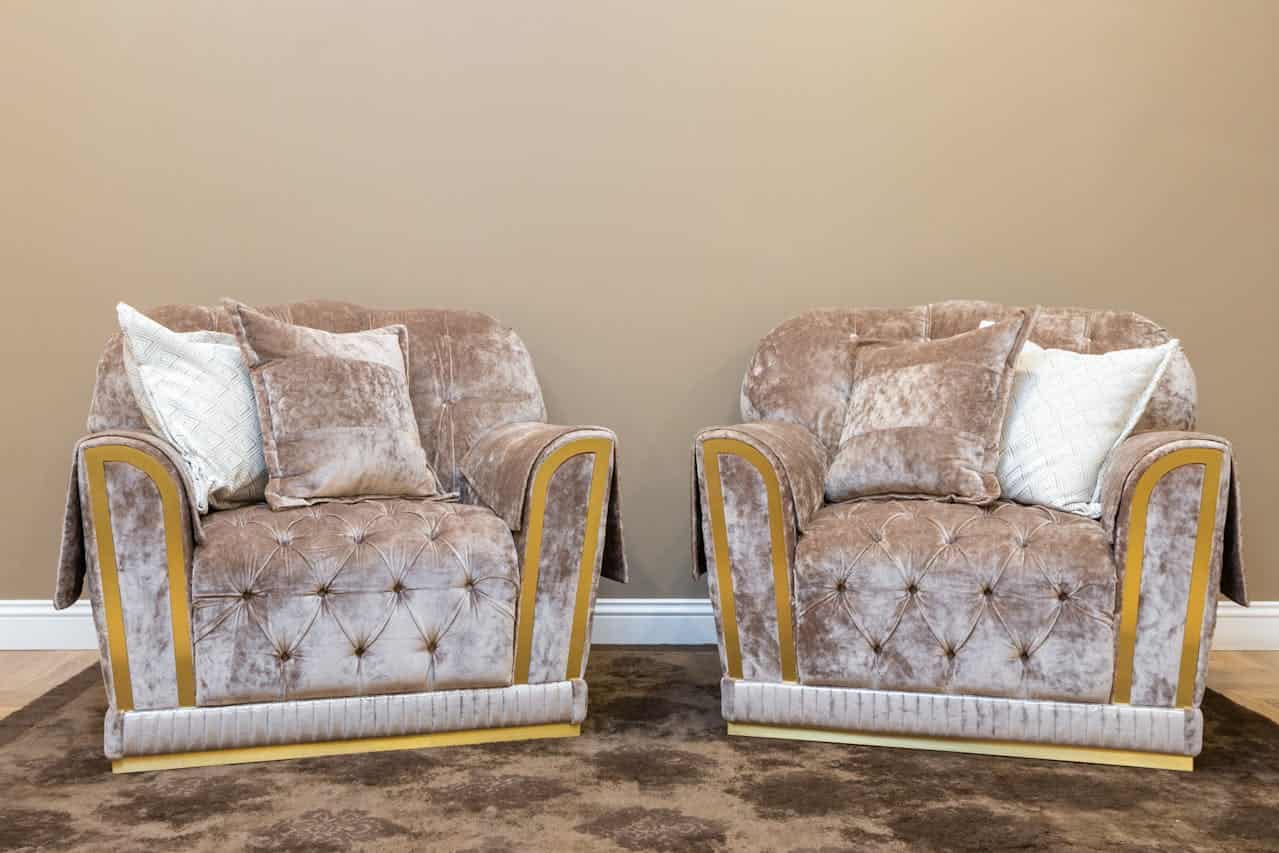 Two luxurious taupe velvet armchairs with tufted cushions, gold trim accents, and light decorative plush velvet throw pillows against beige wall