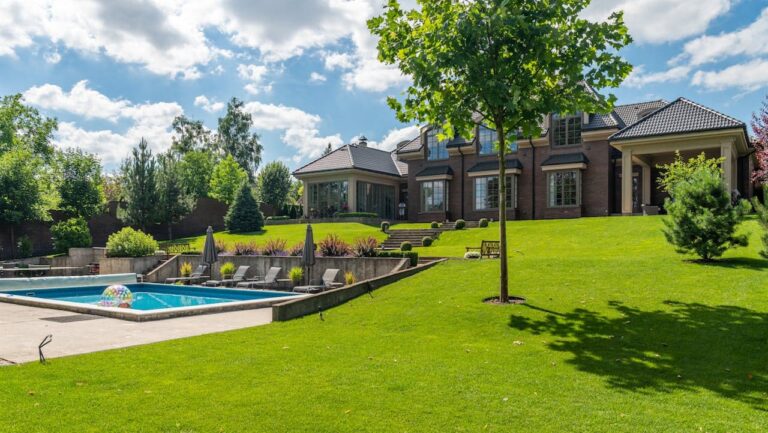 Large brick mansion with black roof on manicured lawn with swimming pool, lounge chairs, and surrounding trees