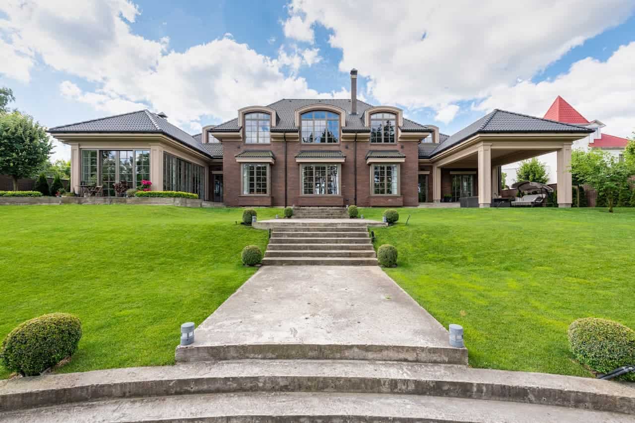 Luxurious brick mansion with dark roof, symmetrical design, manicured lawn, stone pathway, dormer windows, and covered porches on both sides