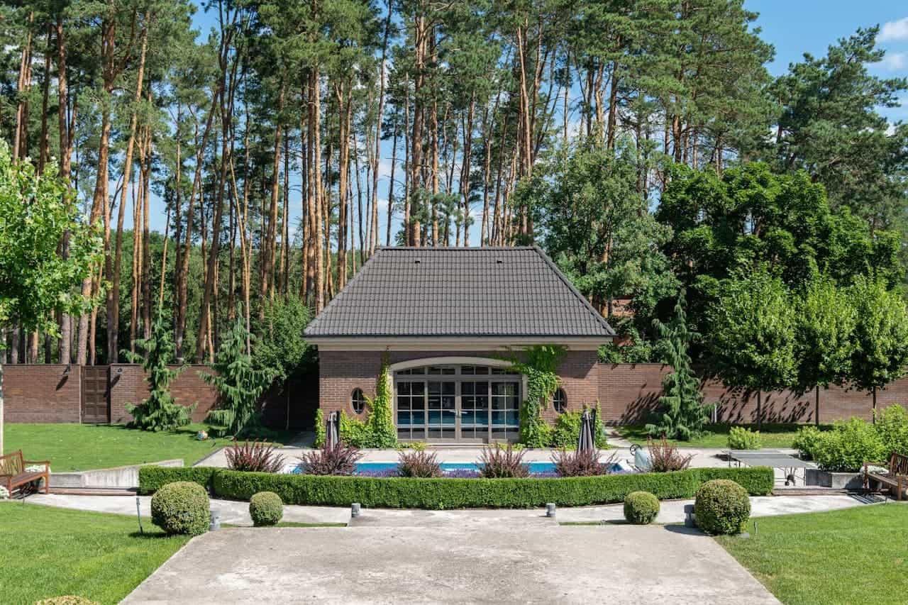 Modern brick pool house with gray roof, surrounded by lush greenery, manicured living hedge borders, concrete path, and tall pine trees