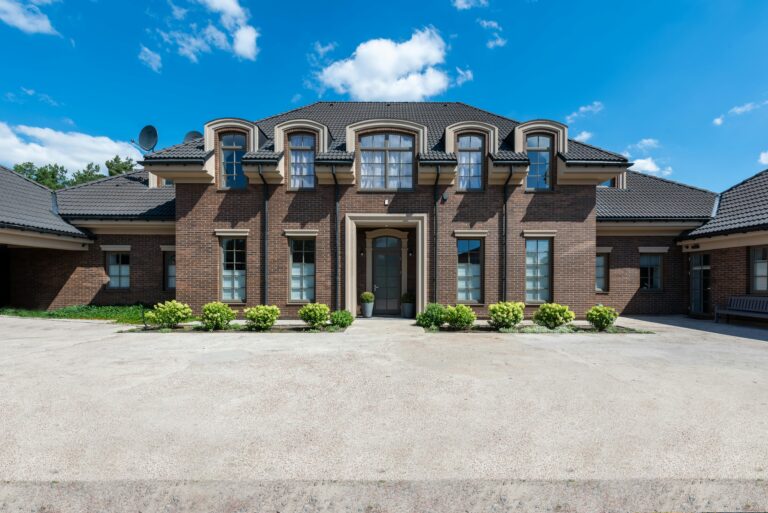 Symmetrical front with six dormer windows, central columned entrance, wide driveway in front, neatly trimmed green shrubs
