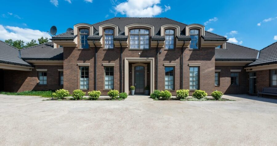 Symmetrical front with six dormer windows, central columned entrance, wide driveway in front, neatly trimmed green shrubs