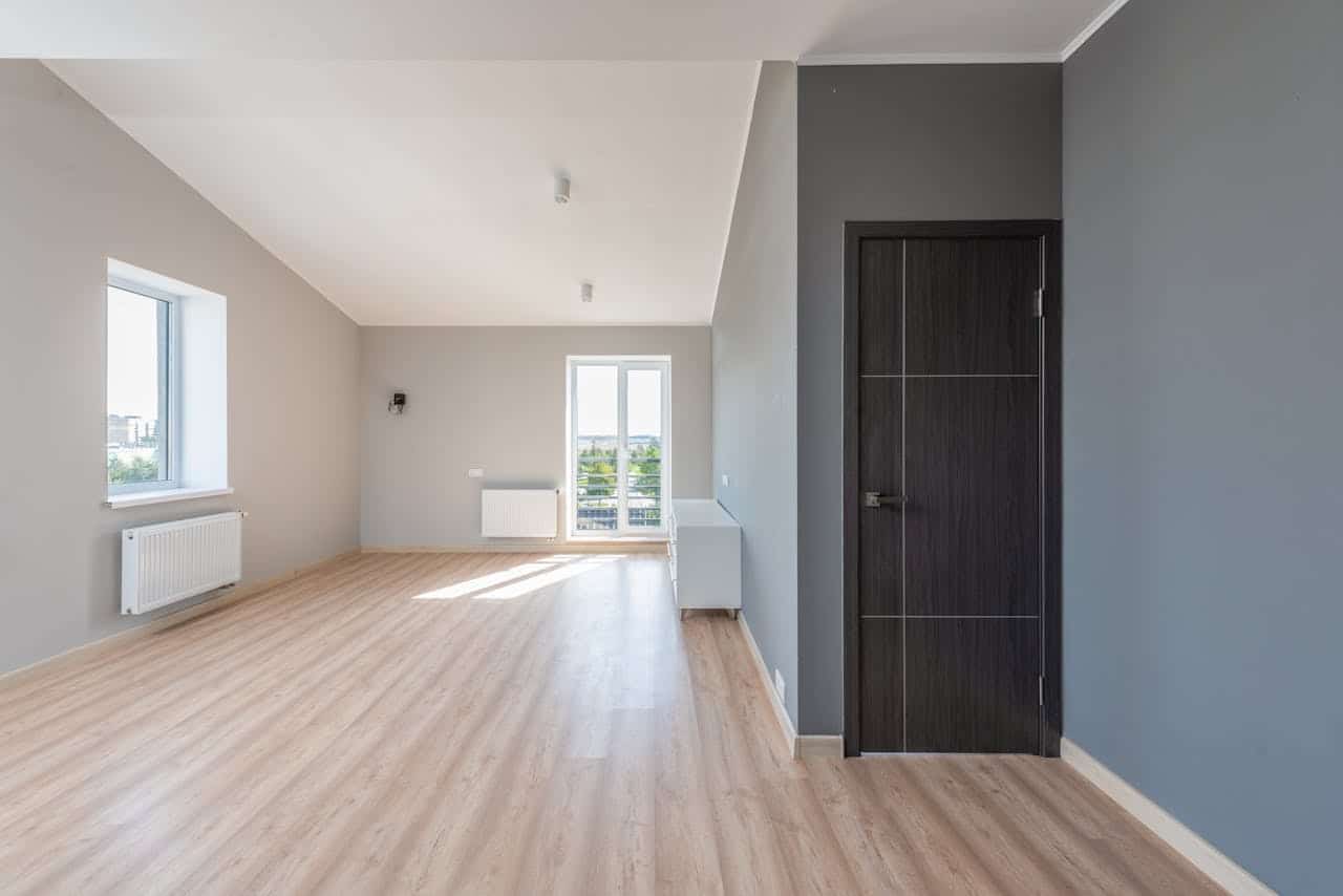 Empty modern apartment with vinyl flooring, gray walls, white ceiling, dark door, windows with city view, and radiators