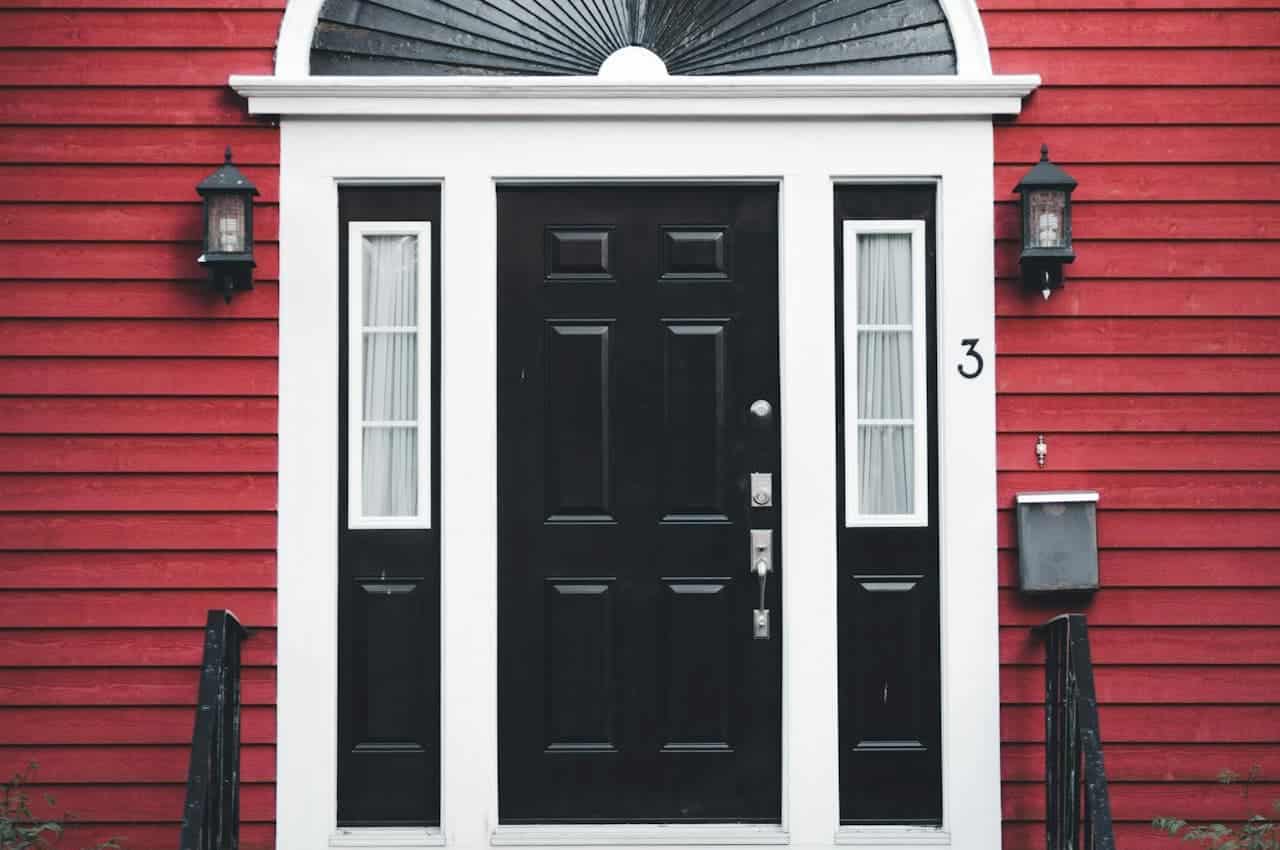 A black front door with two side windows, surrounded by white trim, located on a red wooden house, featuring two black lantern-style lights, a mailbox on the side, and the number "3" displayed on the door