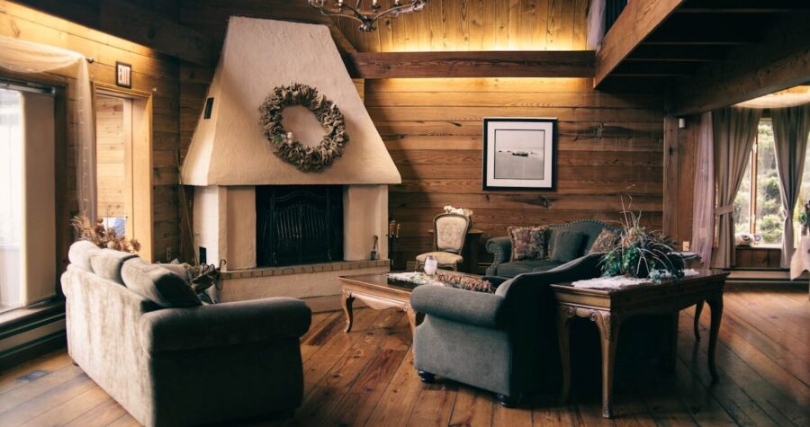 Cozy rustic cabin living room with wooden walls, white fireplace, chandelier, comfortable sofas, and natural light