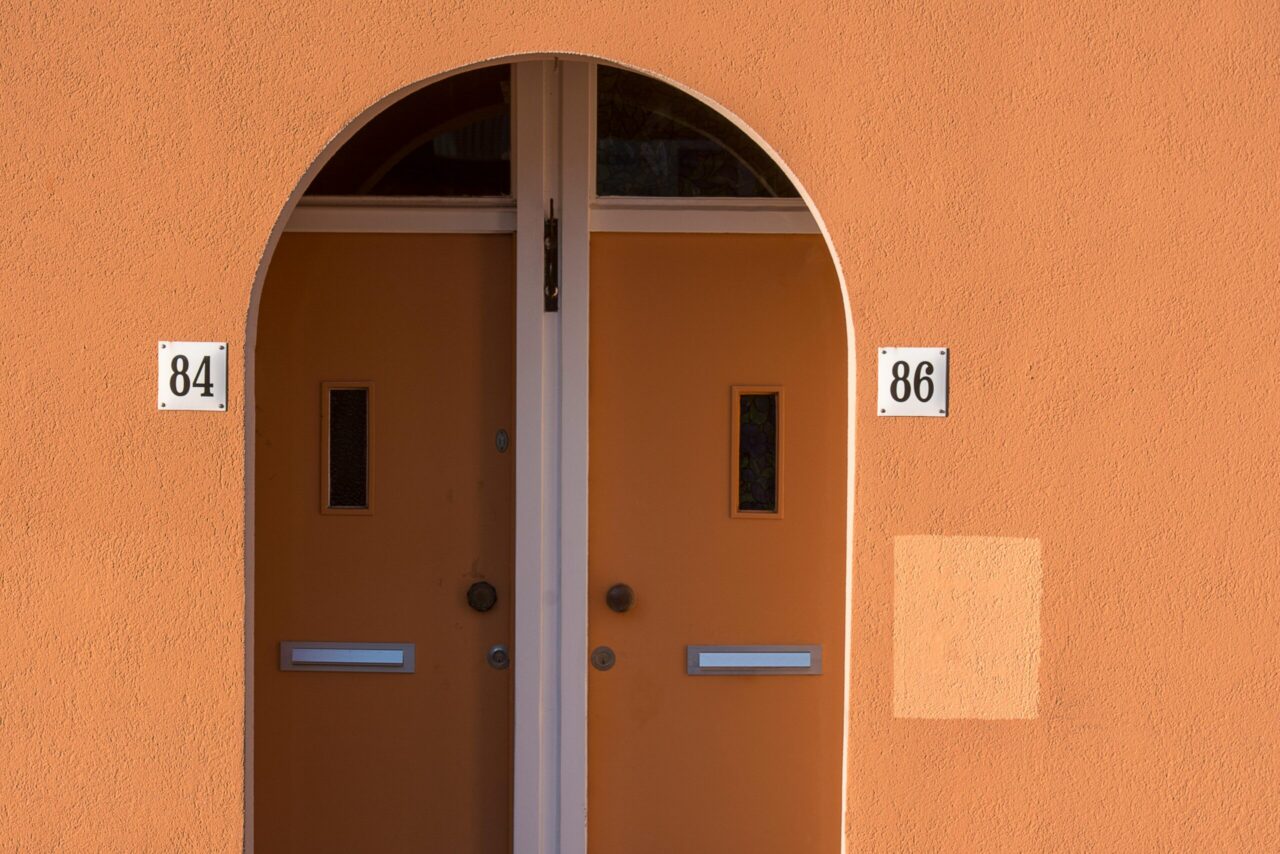 Two orange doors labeled 84 and 86, under an arched entryway, with mail slots and small windows, set in a smooth orange wall
