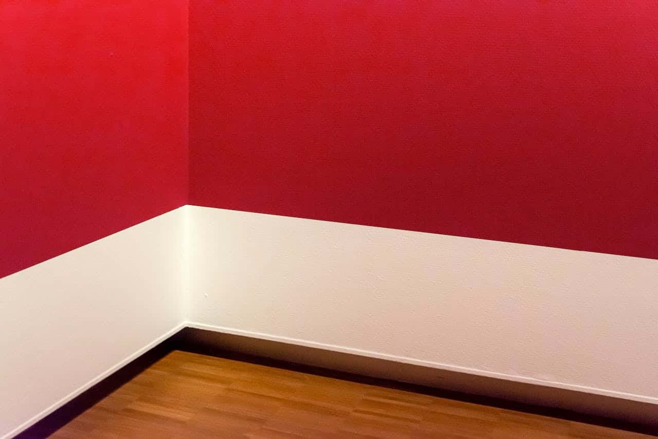Empty room corner with bright red upper walls, white lower walls, and wooden floor with dark baseboard trim