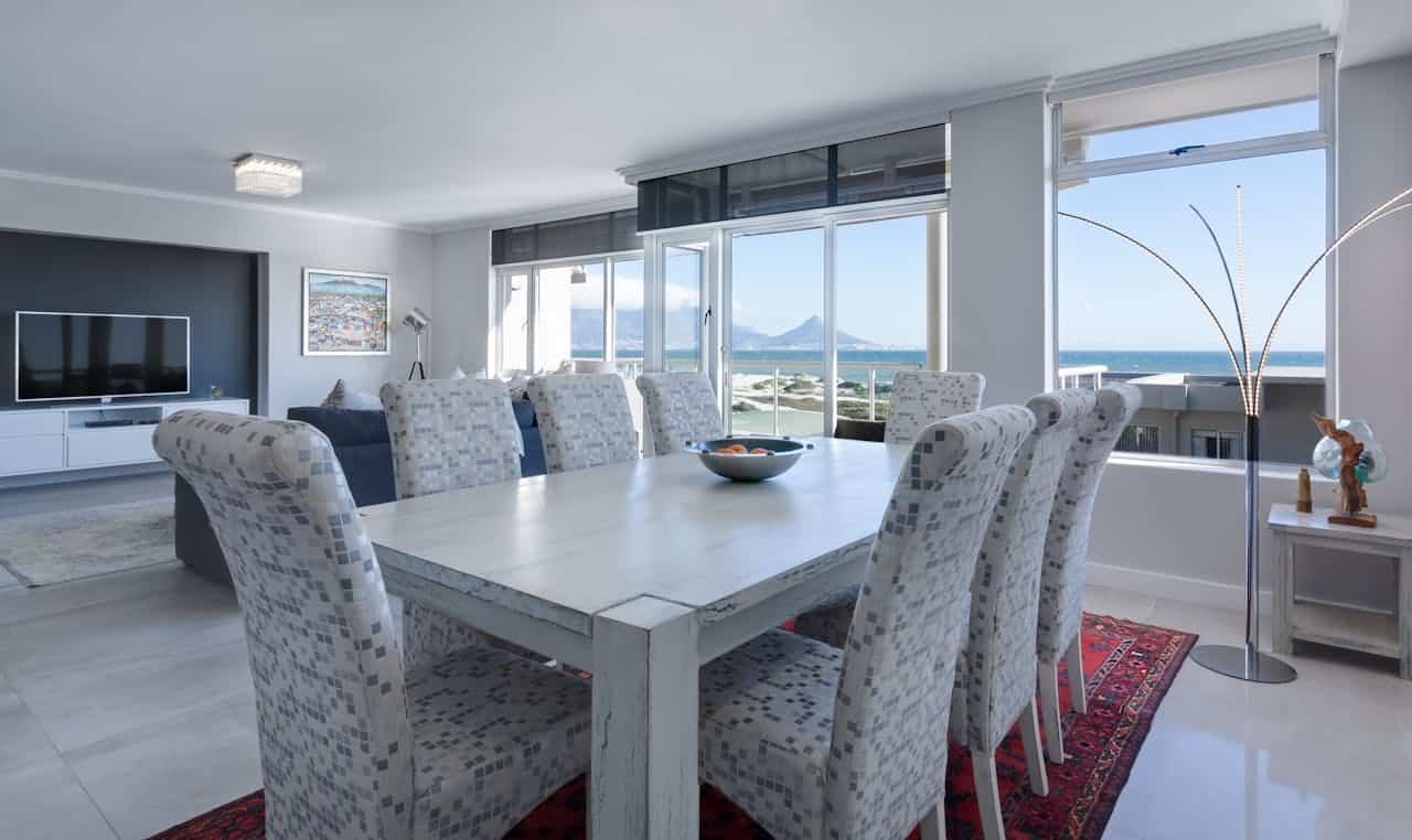 Modern coastal dining area with white table, patterned chairs, ocean view windows, adjacent living room, and red area rug