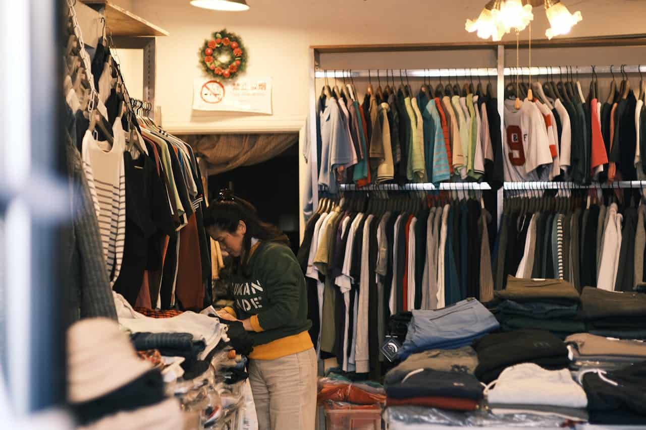 Person in green sweatshirt organizing merchandise in clothing store with hanging garments, folded stacks, and holiday wreath