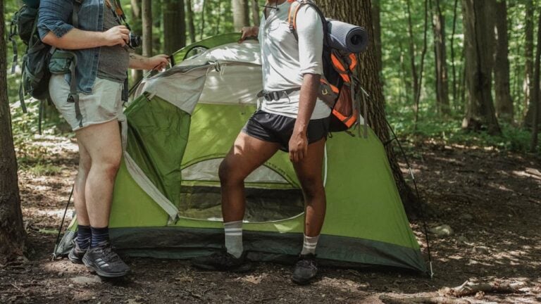 Two hikers setting up green tent in forest, one holding camera while other adjusts gear with backpacks nearby