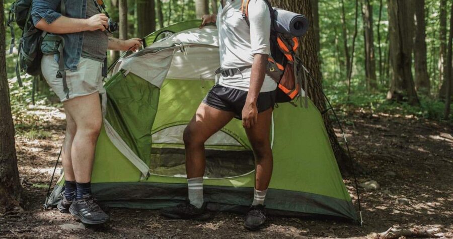 Two hikers setting up green tent in forest, one holding camera while other adjusts gear with backpacks nearby