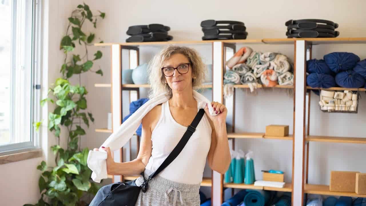 Middle-aged woman in a yoga studio, wearing glasses and casual workout clothes, holding a gym bag and towel, standing in front of shelves with yoga mats, bolsters, and props, natural light from window, green plant in the corner