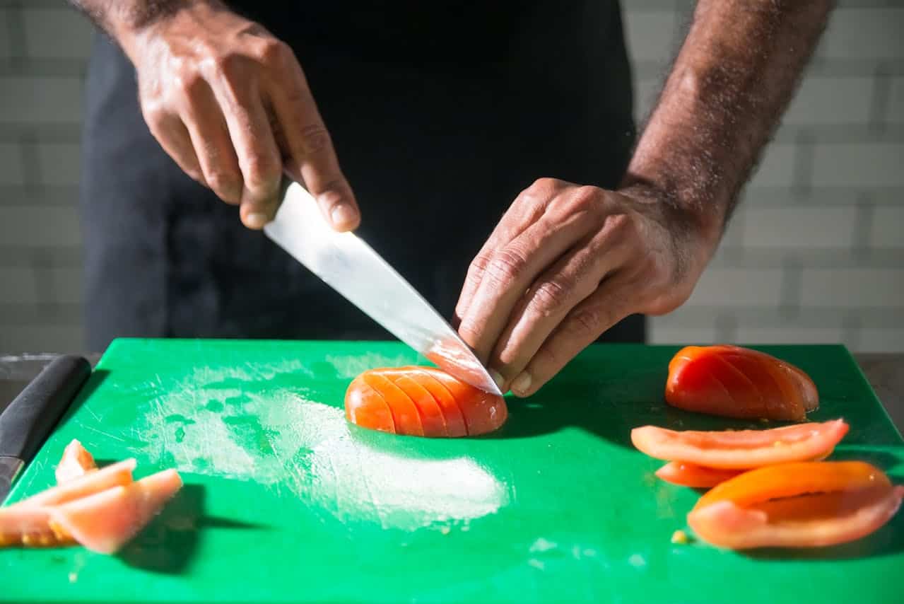 Person slicing a ripe tomato with a sharp knife on a green cutting board, hands positioned for precision, preparing ingredients for cooking or meal preparation in a kitchen setting