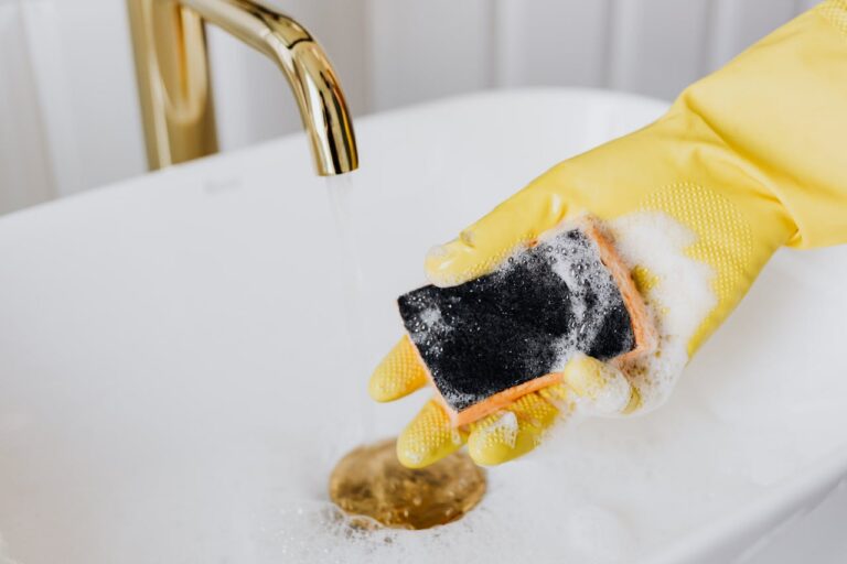 Hand wearing yellow rubber glove holding soapy black sponge while cleaning white sink with gold faucet visible