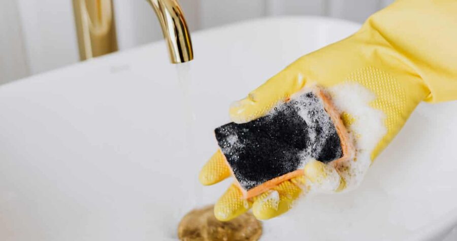 Hand wearing yellow rubber glove holding soapy black sponge while cleaning white sink with gold faucet visible