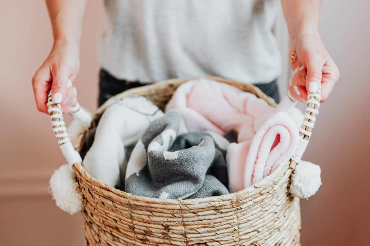 Hands holding woven basket with soft blankets in pink, white, and gray against a blush pink wall