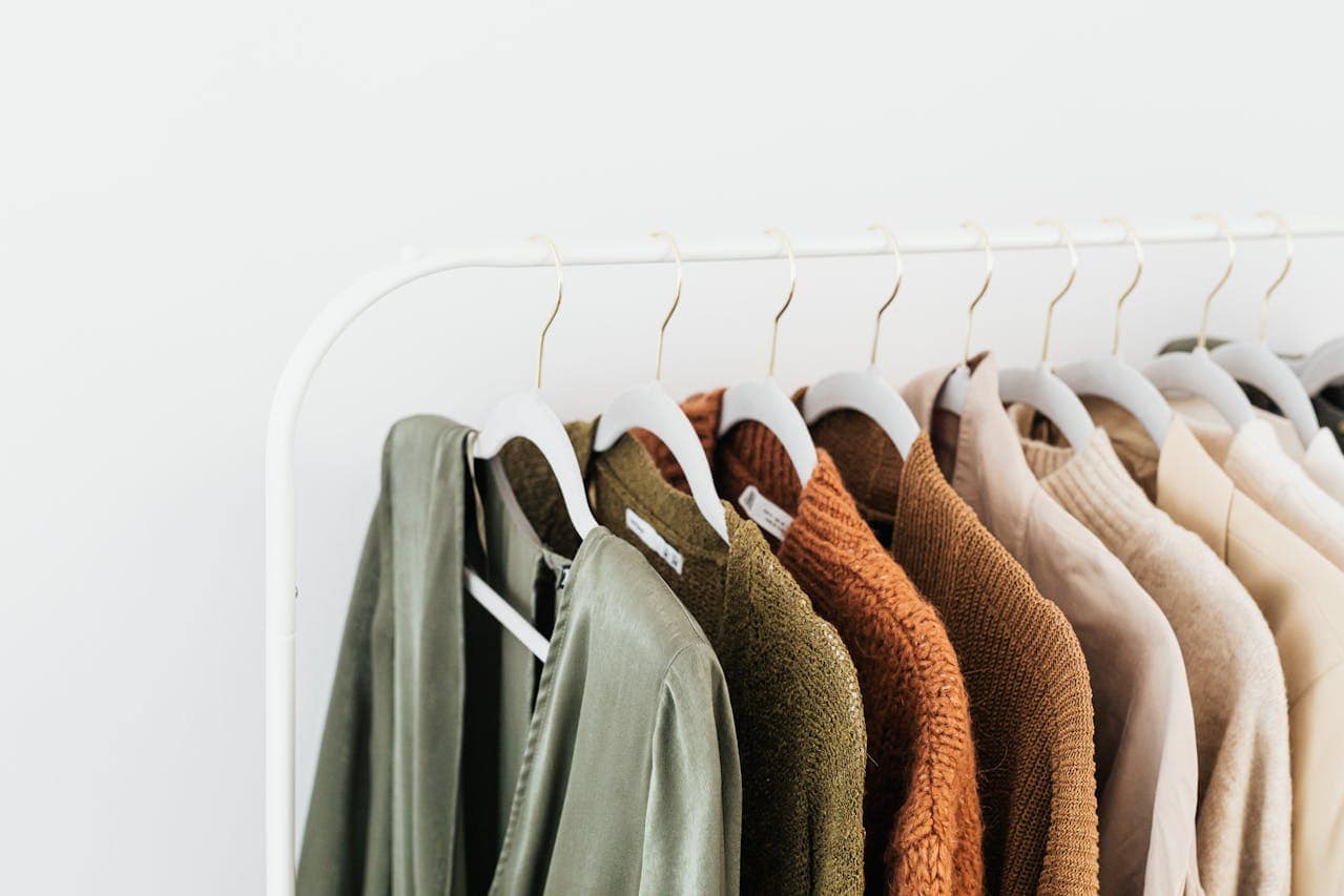 Minimalist white clothing rack with earth-toned garments on matching white hangers, arranged from sage green to cream colors