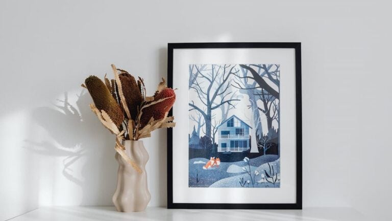 Framed artwork showing blue winter house scene with fox displayed beside cream ceramic vase containing dried flowers