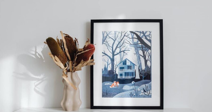 Framed artwork showing blue winter house scene with fox displayed beside cream ceramic vase containing dried flowers