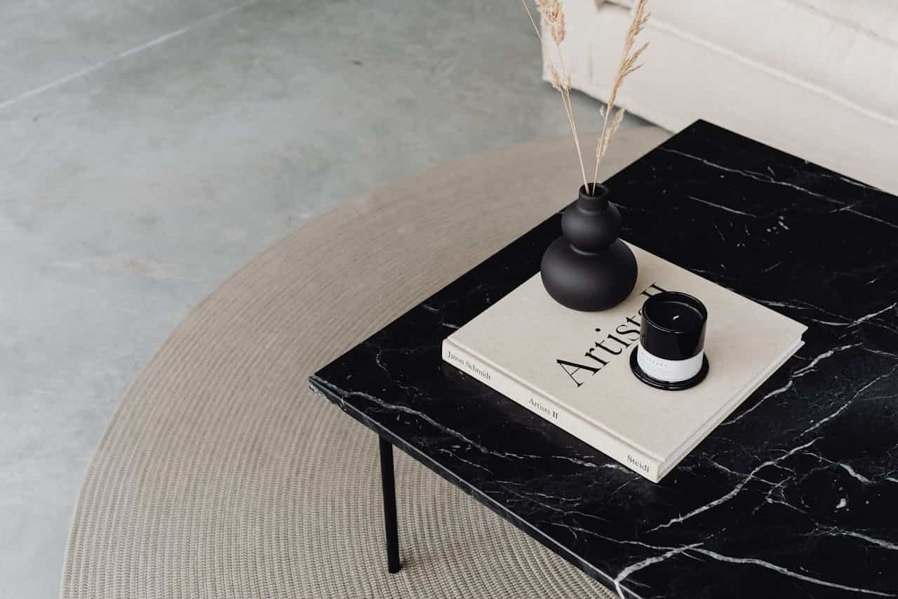 Minimalist black marble coffee table with art book, matte black vase containing dried grass, and candle on round beige rug