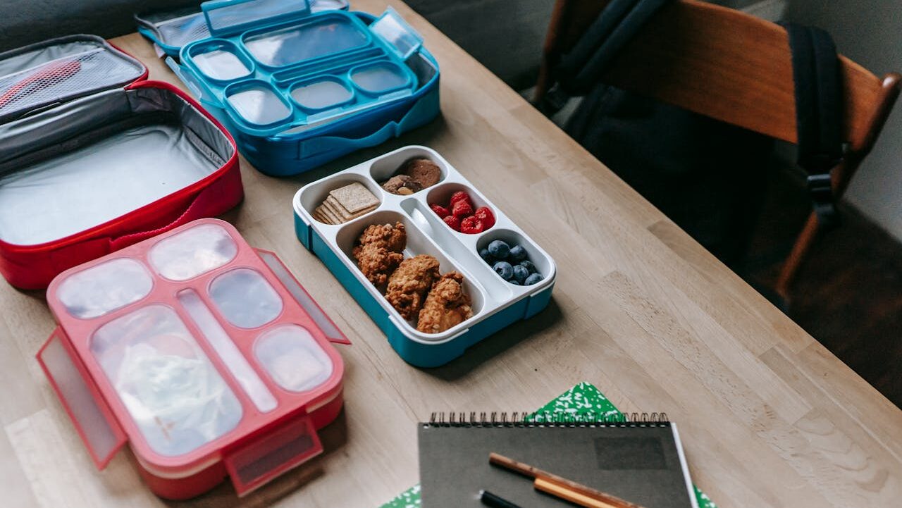 Wooden table with soft-sided lunchboxes containing cookies, berries, and crackers, plus notebooks and pencils for school or work