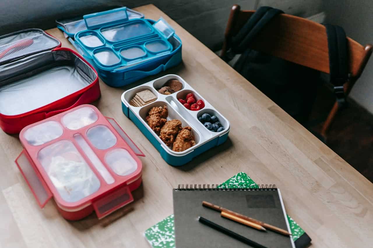 Wooden table with soft-sided lunchboxes containing cookies, berries, and crackers, plus notebooks and pencils for school or work