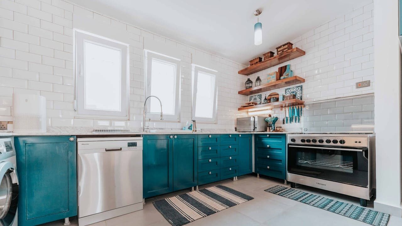 Modern kitchen with teal lower cabinets, white subway tile walls, wooden open shelving, stainless appliances, striped rugs, and three windows