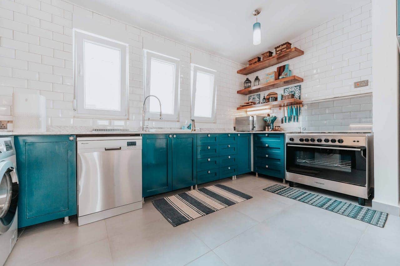 Modern kitchen with teal lower cabinets, white subway tile walls, wooden open shelving, stainless appliances, striped rugs, and three windows