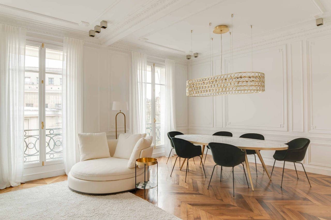 Elegant white dining room with oval table, black chairs, gold chandelier, herringbone floors, round sofa, and French windows