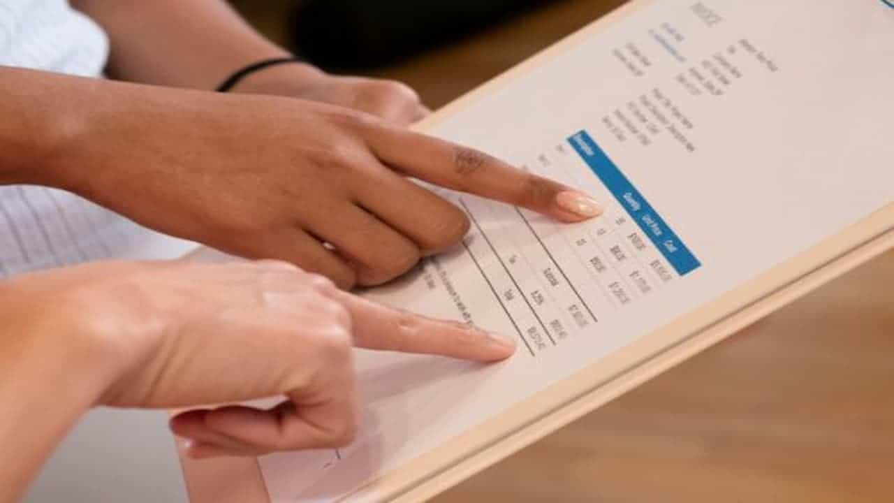 Two people pointing at printed invoice on clipboard, close-up of hands, visible cost breakdown with itemized table, discussing billing details or purchase agreement