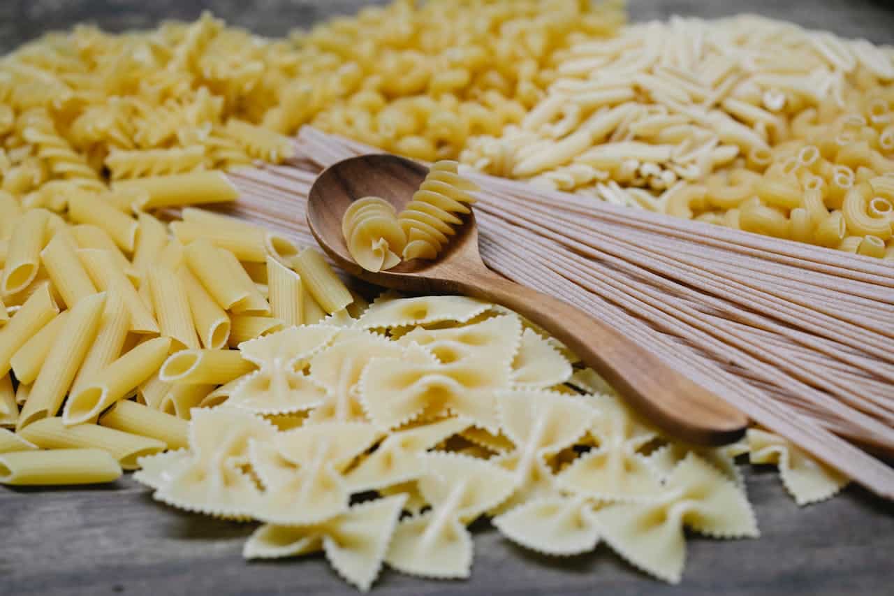 Assorted uncooked pasta types including penne, farfalle, fusilli, macaroni, and spaghetti, arranged on a wooden surface, with a wooden spoon holding a few fusilli pieces, showcasing variety and texture for culinary preparation or food display