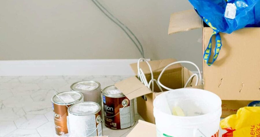 Several cans of wall paint, a white plastic bucket with a stirrer, cardboard boxes, and electrical cables arranged on a tiled floor in front of a freshly painted wall
