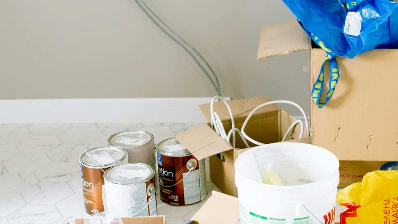 Several cans of wall paint, a white plastic bucket with a stirrer, cardboard boxes, and electrical cables arranged on a tiled floor in front of a freshly painted wall