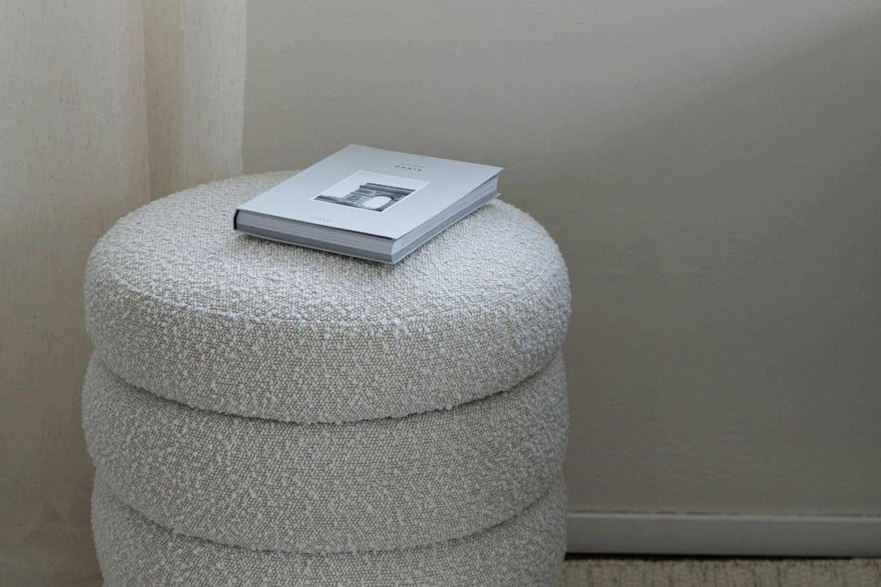 White hardcover book resting on round cream-colored boucle fabrics ottoman against light gray wall in minimalist corner