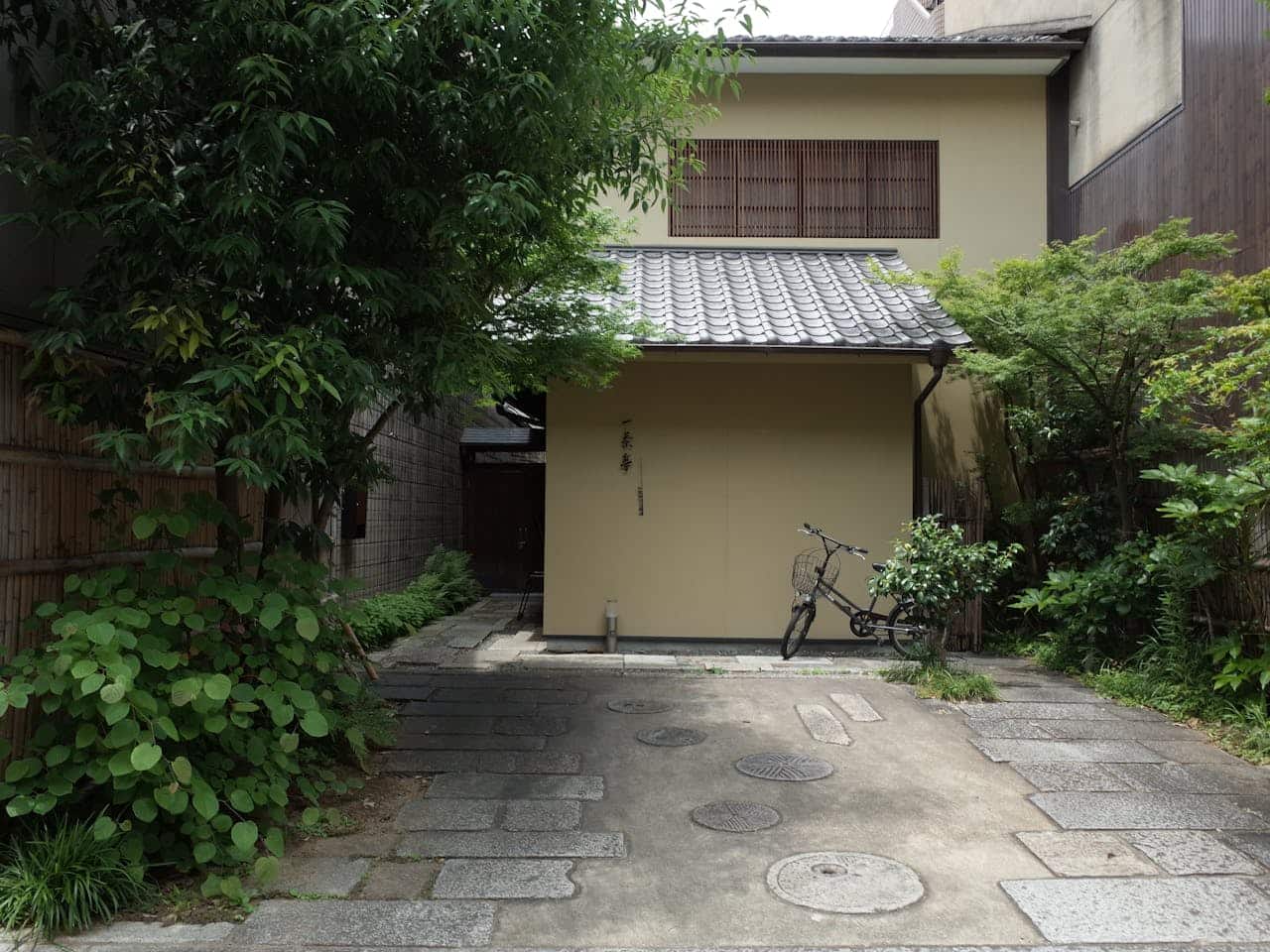 Traditional Japanese house with tiled roof, cream walls, wooden lattice window, surrounded by lush greenery, bicycle parked outside entrance