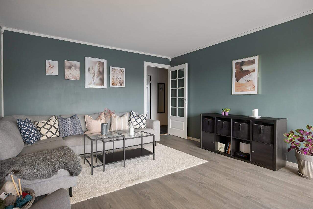 Modern living room with sage green walls, gray sectional sofa, chequered prints on throw pillows, glass coffee tables, framed artwork, and black storage unit