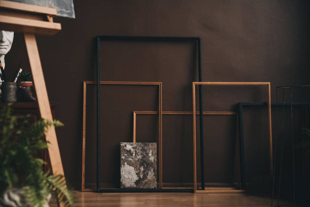 The image shows a set of empty picture frames stacked against a wall, with a piece of artwork partially visible at the bottom, an easel in the background, potted plants nearby, and a neutral brown wall behind, creating a calm, artistic atmosphere