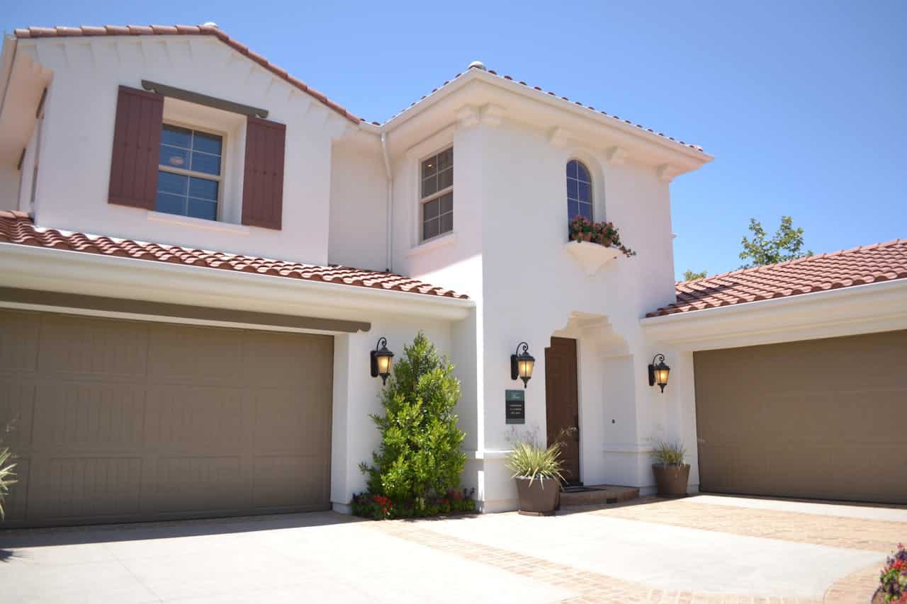 Mediterranean-style white stucco house with terracotta roof tiles, brown shutters, two garage doors, outdoor lighting, and potted plants