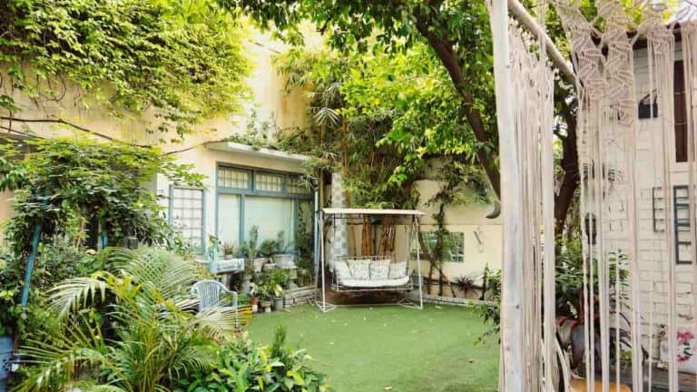 Cozy, multi-purpose outdoor space with lush green plants, a swing bench, hanging macrame, and potted plants arranged around a grassy yard area in front of a home
