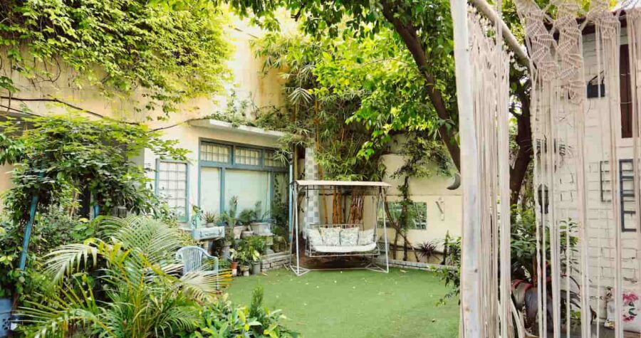 Cozy, multi-purpose outdoor space with lush green plants, a swing bench, hanging macrame, and potted plants arranged around a grassy yard area in front of a home