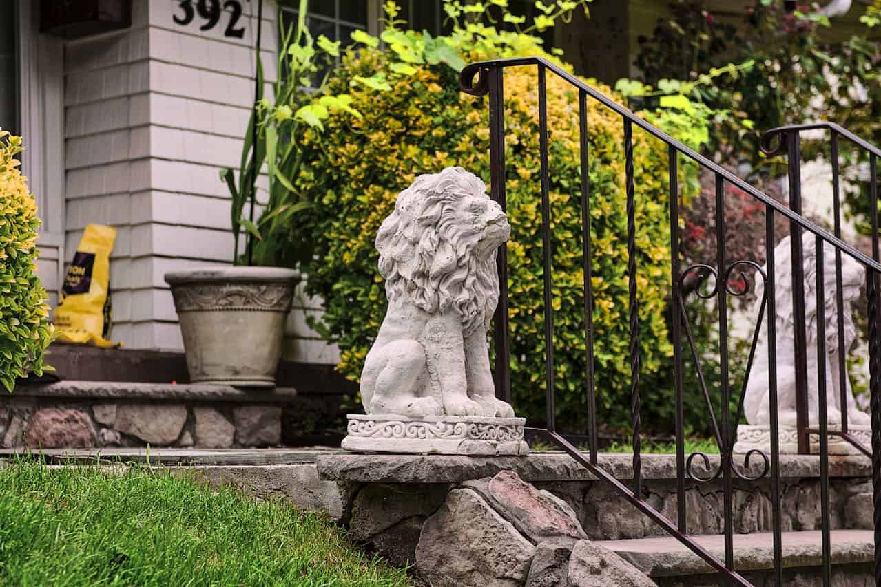Stone lion stone statue sits on rocky steps near house, with decorative metal railing, potted plants, and yellow-green foliage