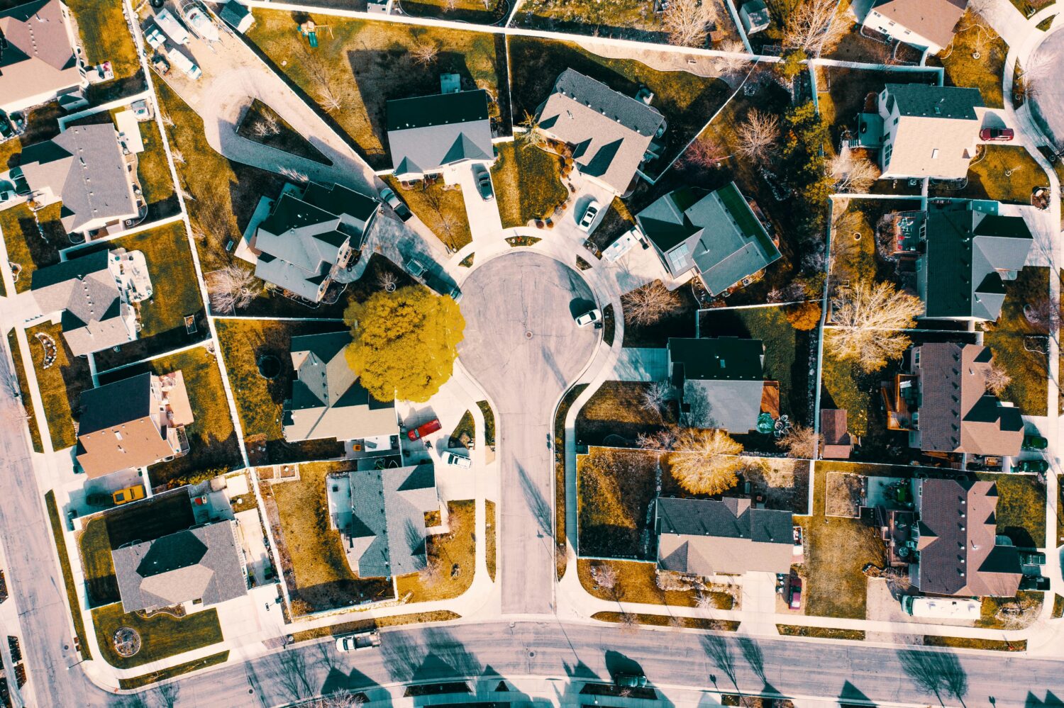 Aerial view of a suburban neighborhood, houses arranged around a cul-de-sac, parked cars along driveways and roads, visible backyards and fences, tree-lined yards with patchy grass, intersecting streets forming a grid pattern