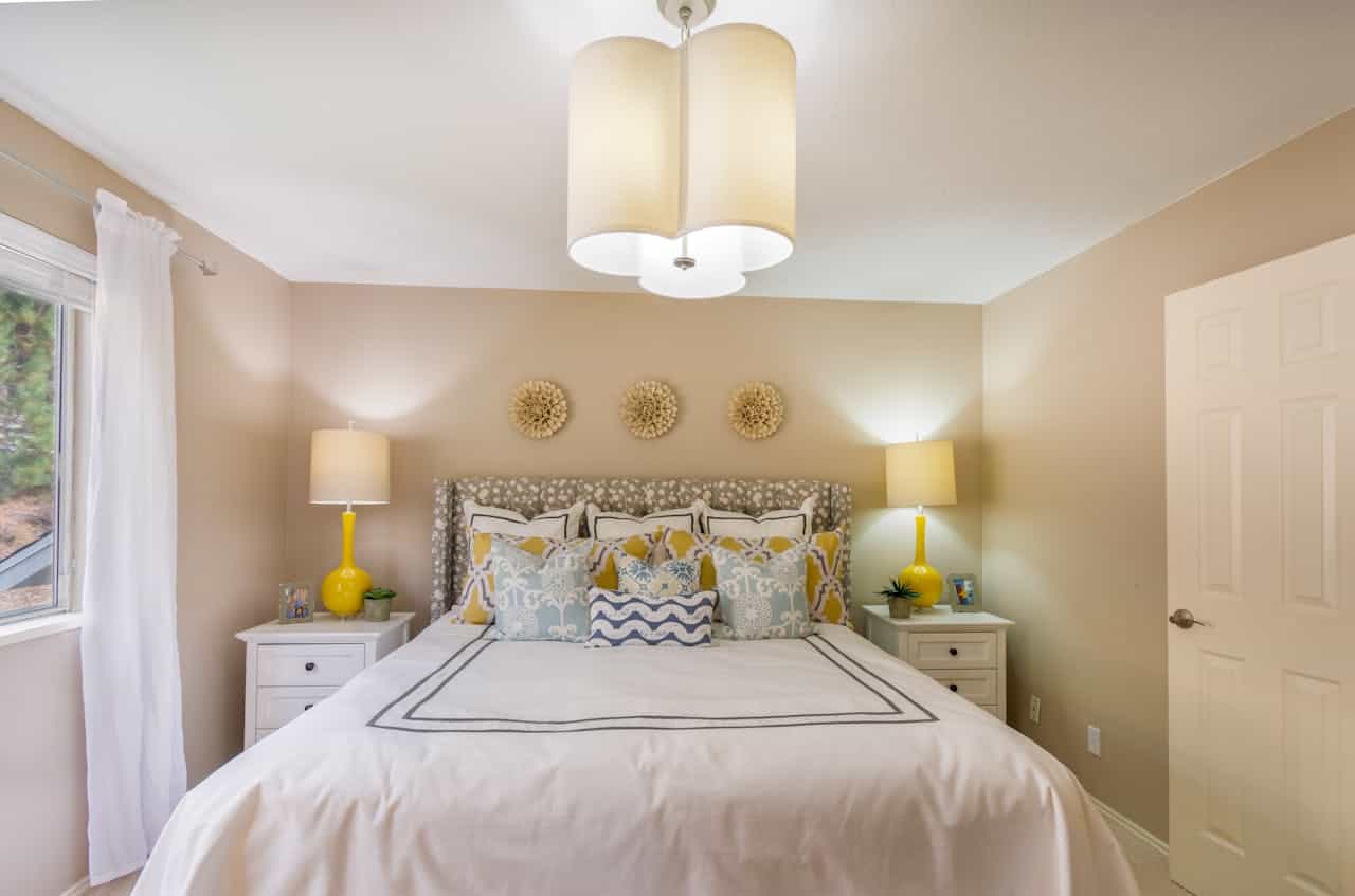Neutral bedroom with white bedding, patterned headboard, yellow accent lamps, decorative pillows, and three sunburst wall decorations