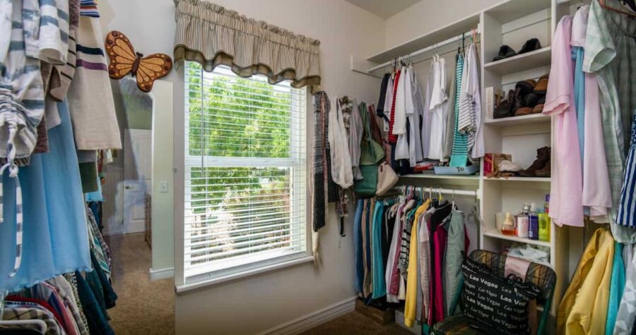 Walk-in closet with white built-in shelving, clothes hanging on rods, window with blinds, butterfly decoration, and carpeted floor