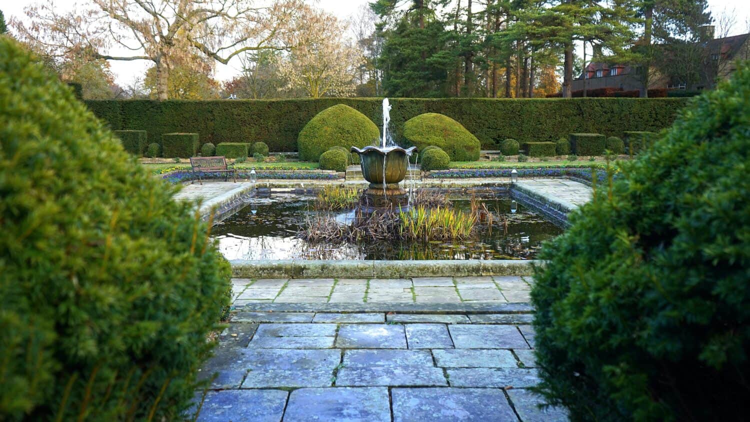 Stone fountain in the center of a formal garden, surrounded by a trimmed hedge, reflective pond, stone paths, and manicured shrubs