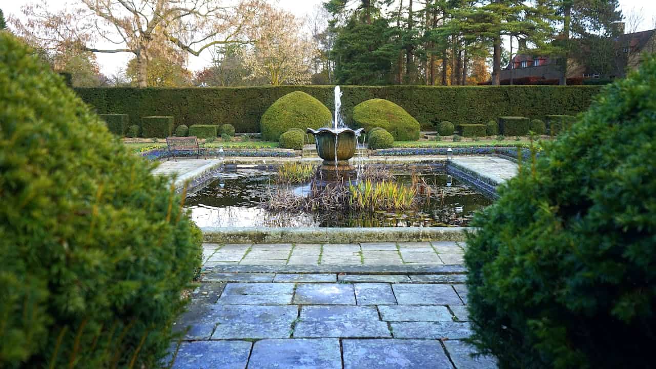 Formal garden with central fountain in stone pond, surrounded by manicured topiary hedges, stone pathway, and tall trees beyond